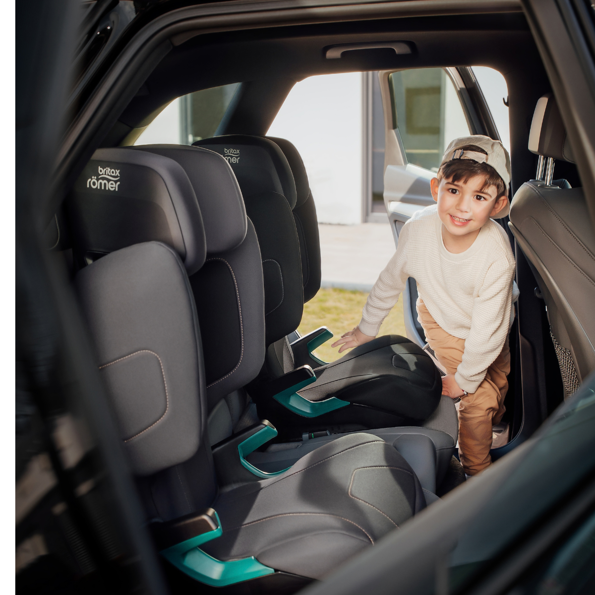 Bambino sorridente accanto ai seggiolini auto Britax R&ouml;mer HI-LINER sul sedile posteriore di un&rsquo;auto.